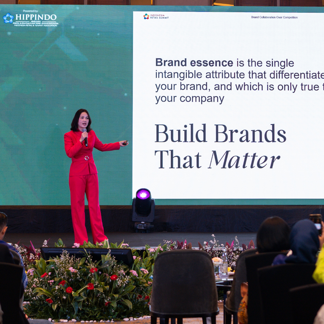 Woman in a red suit presents on a stage beside a large screen displaying 'Build Brands That Matter' at a conference.
